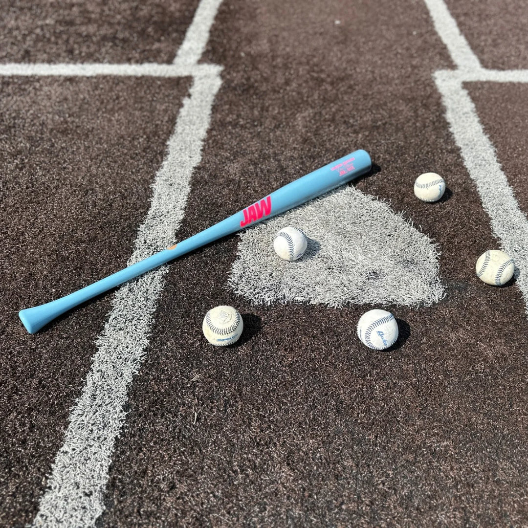 A "cotton candy" colored Jaw Bat laying across home plate with several baseballs scattered around the image. The Jaw Bat letters are bright pink and reflect from the light of the sun.