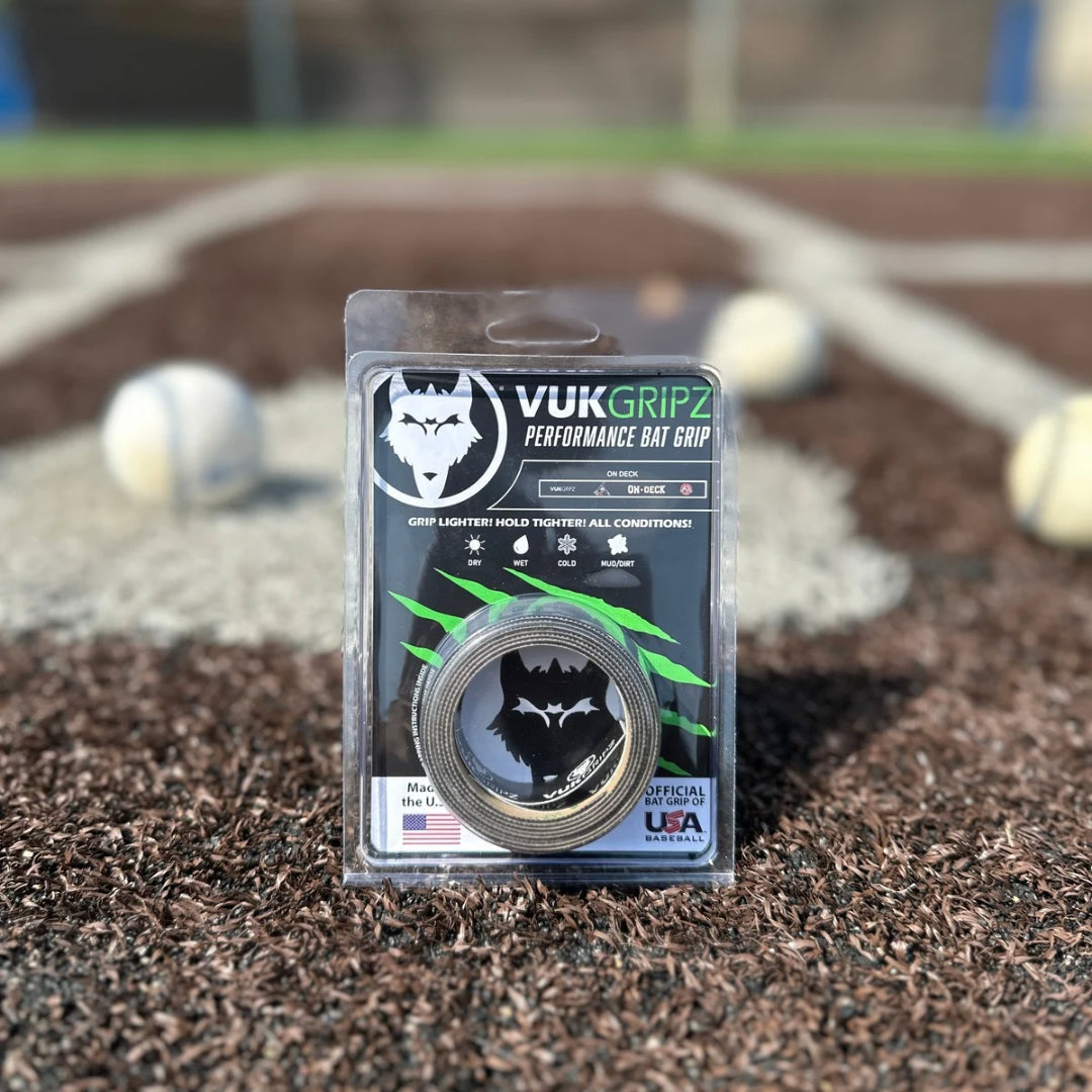 A roll of On-Deck Originals branded VukGripz bat grip tape set upright in front of home plate and a batters box on a sunny day. There are several baseballs in the background