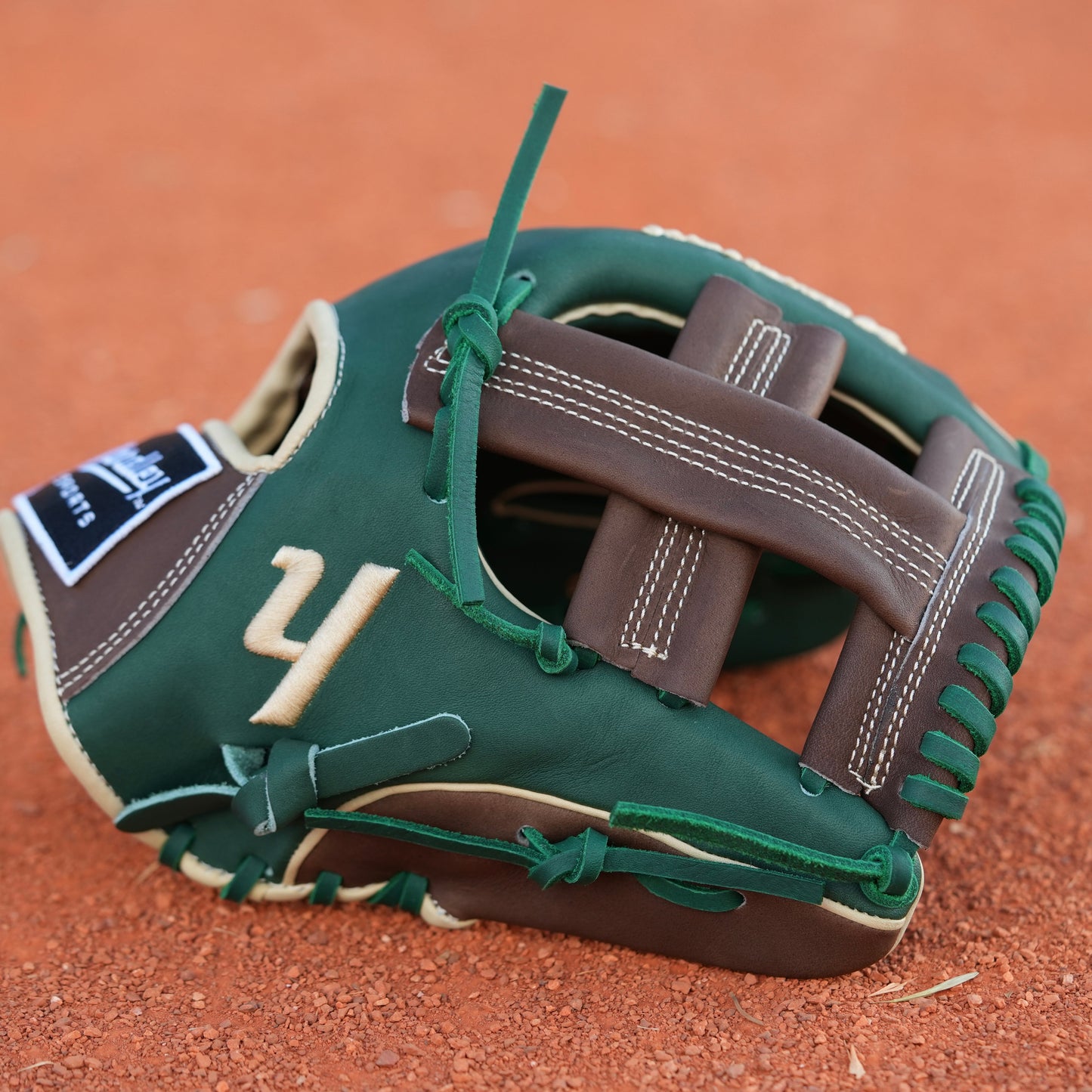 A baseball glove on a dirt field. The glove is primarily green with brown and beige accents, featuring intricate stitching and a patch with text on the wrist area. The letter Y is stitched on the side, indicating Yardley Sports.