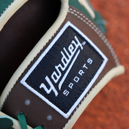 A baseball glove on a dirt field. The glove is primarily green with brown and beige accents, featuring intricate stitching and a patch with text on the wrist area. The letter Y is stitched on the side, indicating Yardley Sports.