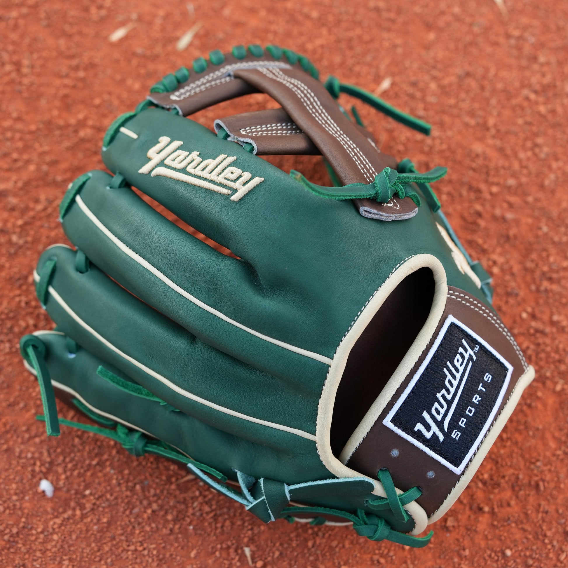 A baseball glove on a dirt field. The glove is primarily green with brown and beige accents, featuring intricate stitching and a patch with text on the wrist area. The letter Y is stitched on the side, indicating Yardley Sports.