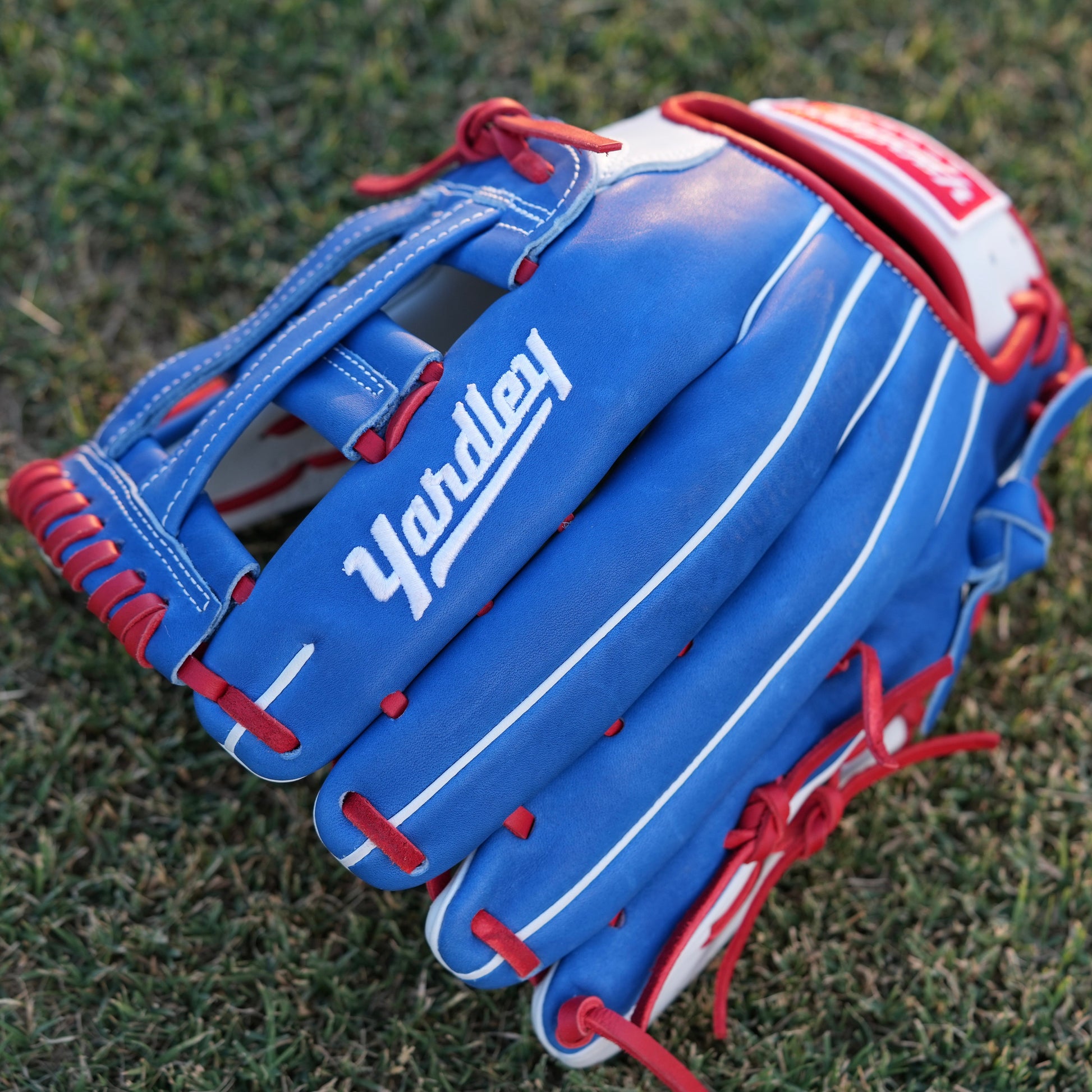  A red Yardley baseball glove with black and white stitching and black laces on a grass field. The glove has a "Y" logo on the thumb area, showcasing its unique design. The webbing is an H-web style