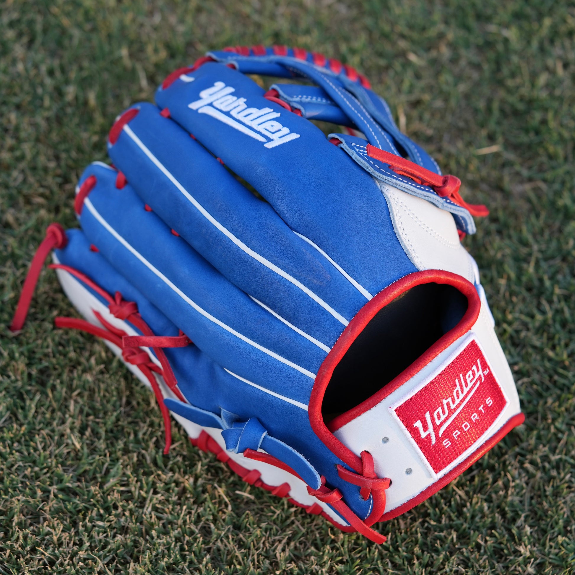  A red Yardley baseball glove with black and white stitching and black laces on a grass field. The glove has a "Y" logo on the thumb area, showcasing its unique design. The webbing is an H-web style