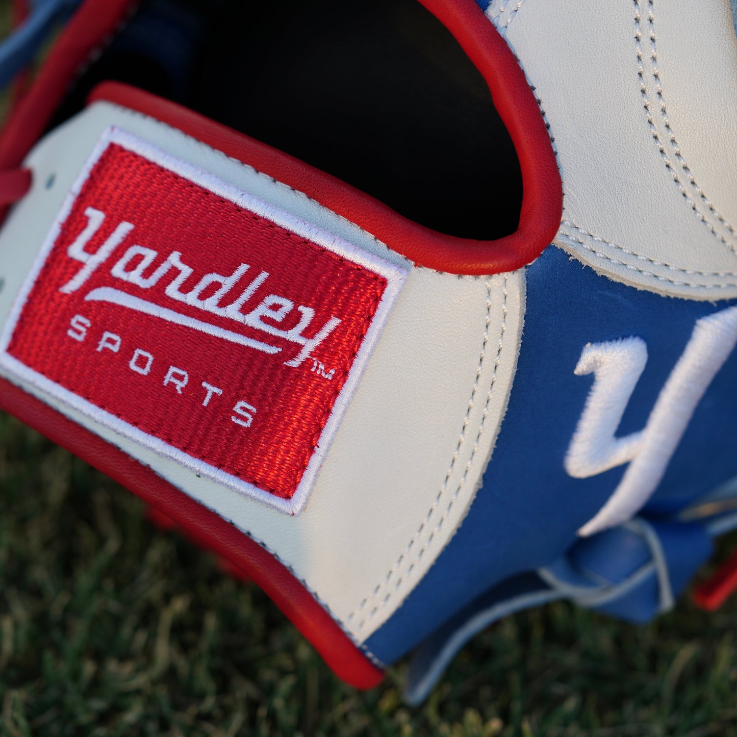  A red Yardley baseball glove with black and white stitching and black laces on a grass field. The glove has a "Y" logo on the thumb area, showcasing its unique design. The webbing is an H-web style