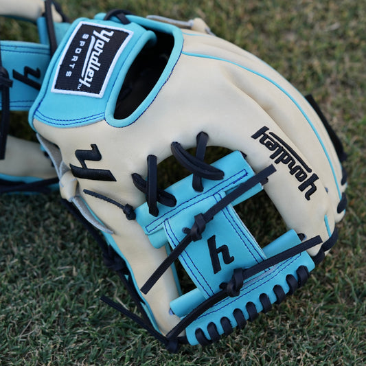 Close-up of a beige baseball glove with light blue accents and black laces, laying on grass. The glove features "Yardley Sports" branding on the wrist strap and "Yardley" on the thumb area. Detailed stitching enhances its high-quality design.