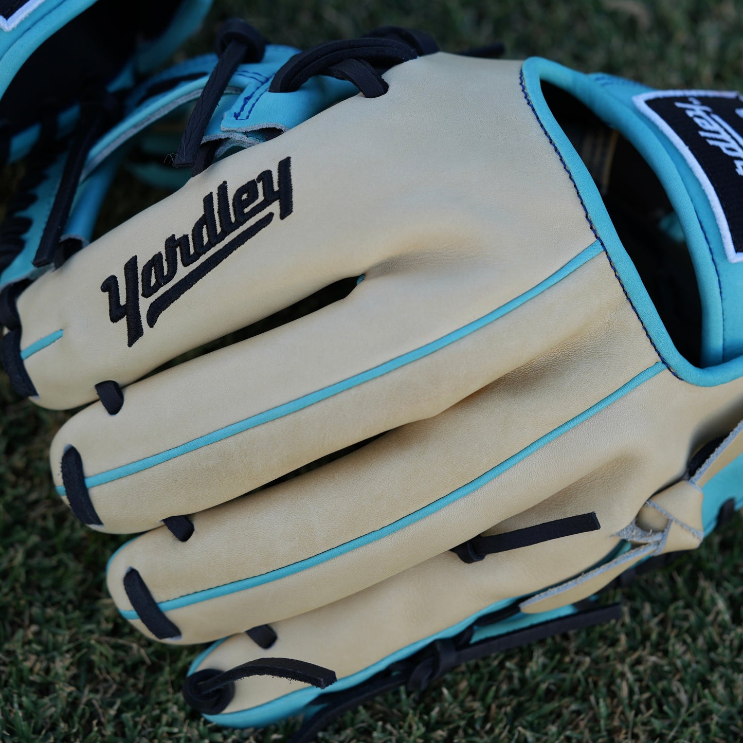 Close-up of a beige baseball glove with light blue accents and black laces, laying on grass. The glove features "Yardley Sports" branding on the wrist strap and "Yardley" on the thumb area. Detailed stitching enhances its high-quality design.