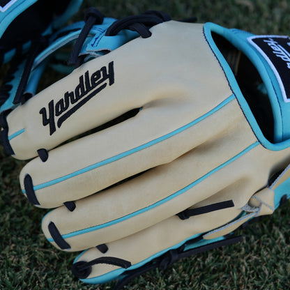 Close-up of a beige baseball glove with light blue accents and black laces, laying on grass. The glove features "Yardley Sports" branding on the wrist strap and "Yardley" on the thumb area. Detailed stitching enhances its high-quality design.