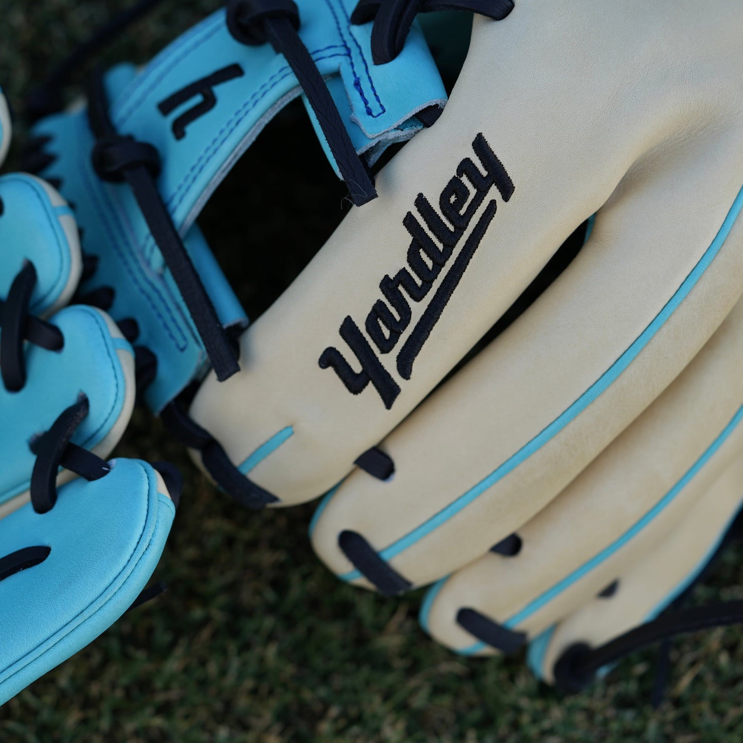 Close-up of a beige baseball glove with light blue accents and black laces, laying on grass. The glove features "Yardley Sports" branding on the wrist strap and "Yardley" on the thumb area. Detailed stitching enhances its high-quality design.