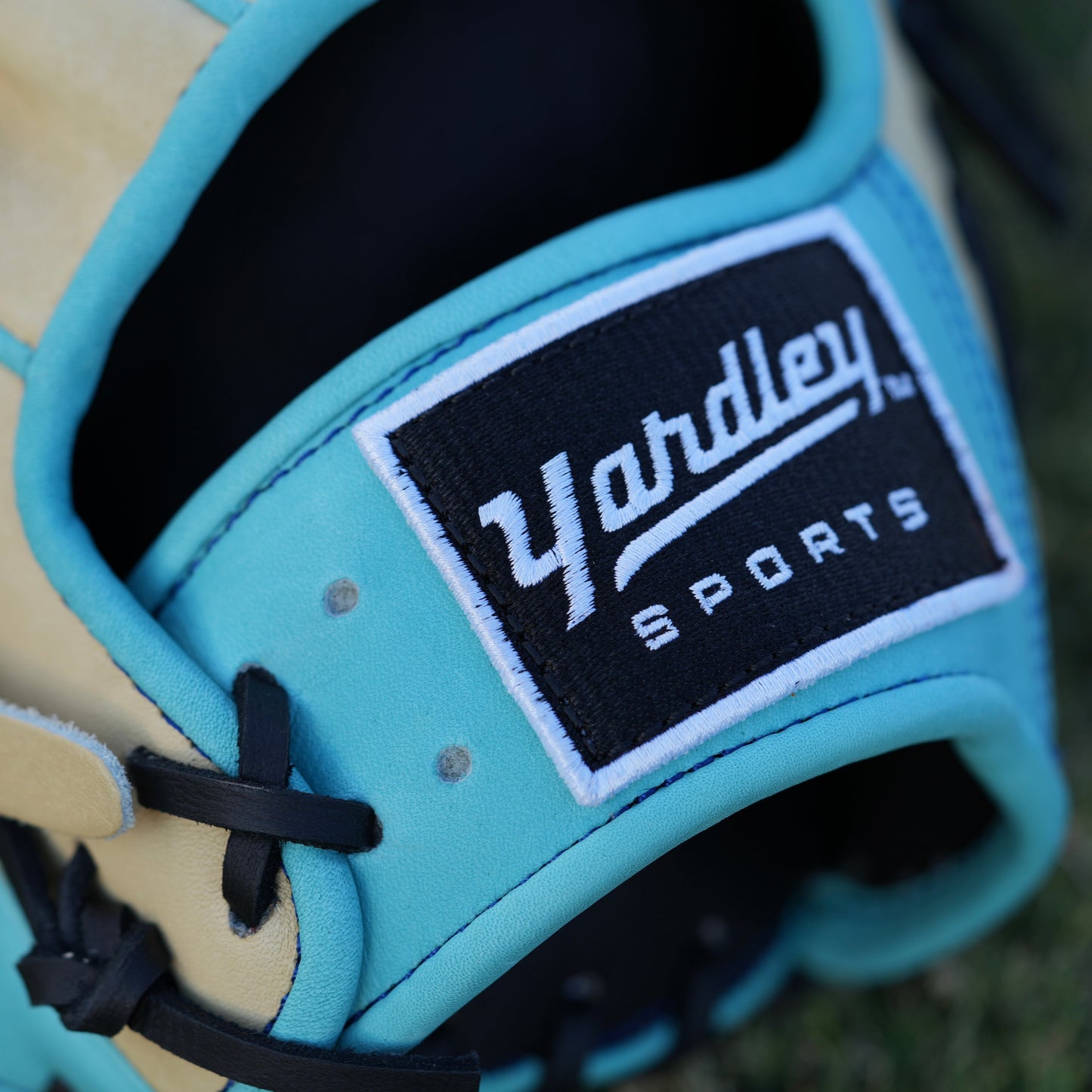 Close-up of a beige baseball glove with light blue accents and black laces, laying on grass. The glove features "Yardley Sports" branding on the wrist strap and "Yardley" on the thumb area. Detailed stitching enhances its high-quality design.