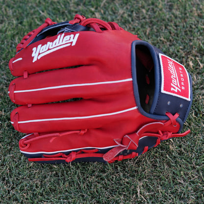 A red and navy blue baseball glove lying on grass, featuring the Yardley Sports logo and branding. The glove has red laces, white embroidered text, including the letter "Y." The design and color scheme highlight the premium quality and craftsmanship associated with Yardley Sports.