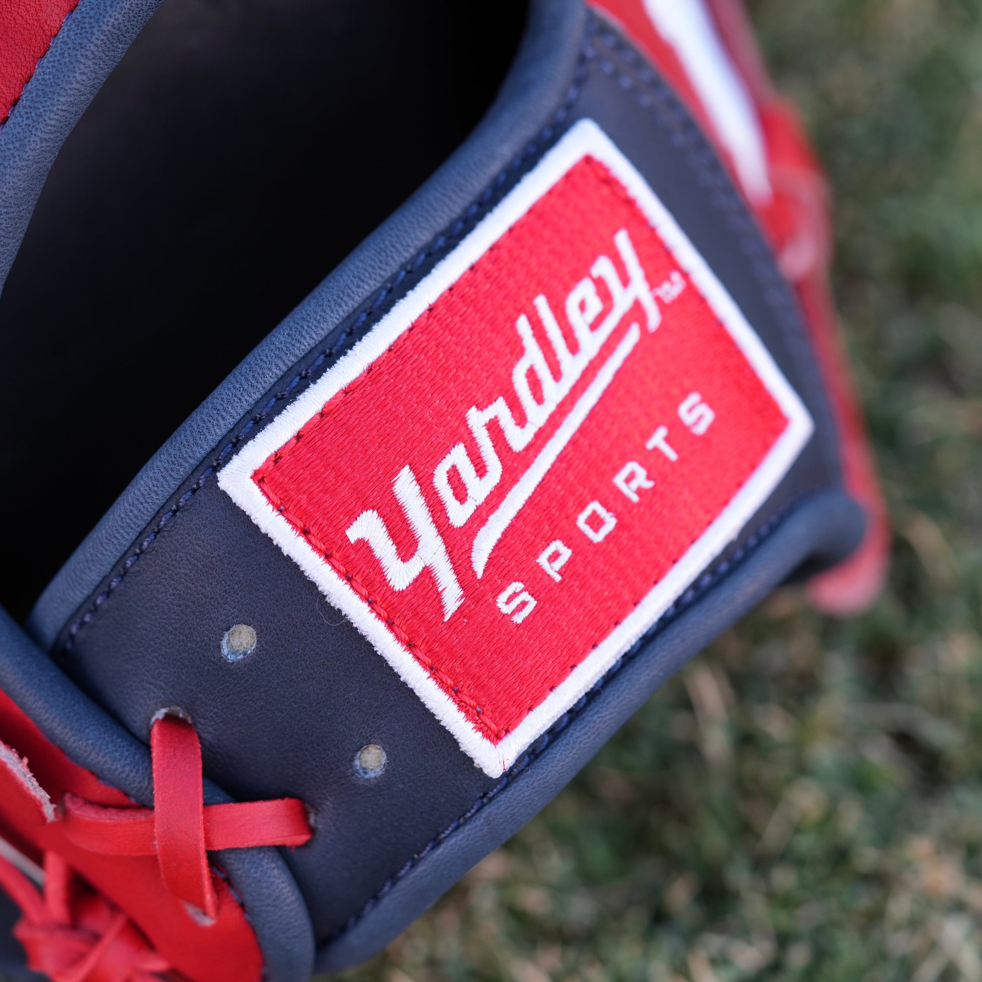 A red and navy blue baseball glove lying on grass, featuring the Yardley Sports logo and branding. The glove has red laces, white embroidered text, including the letter "Y." The design and color scheme highlight the premium quality and craftsmanship associated with Yardley Sports.