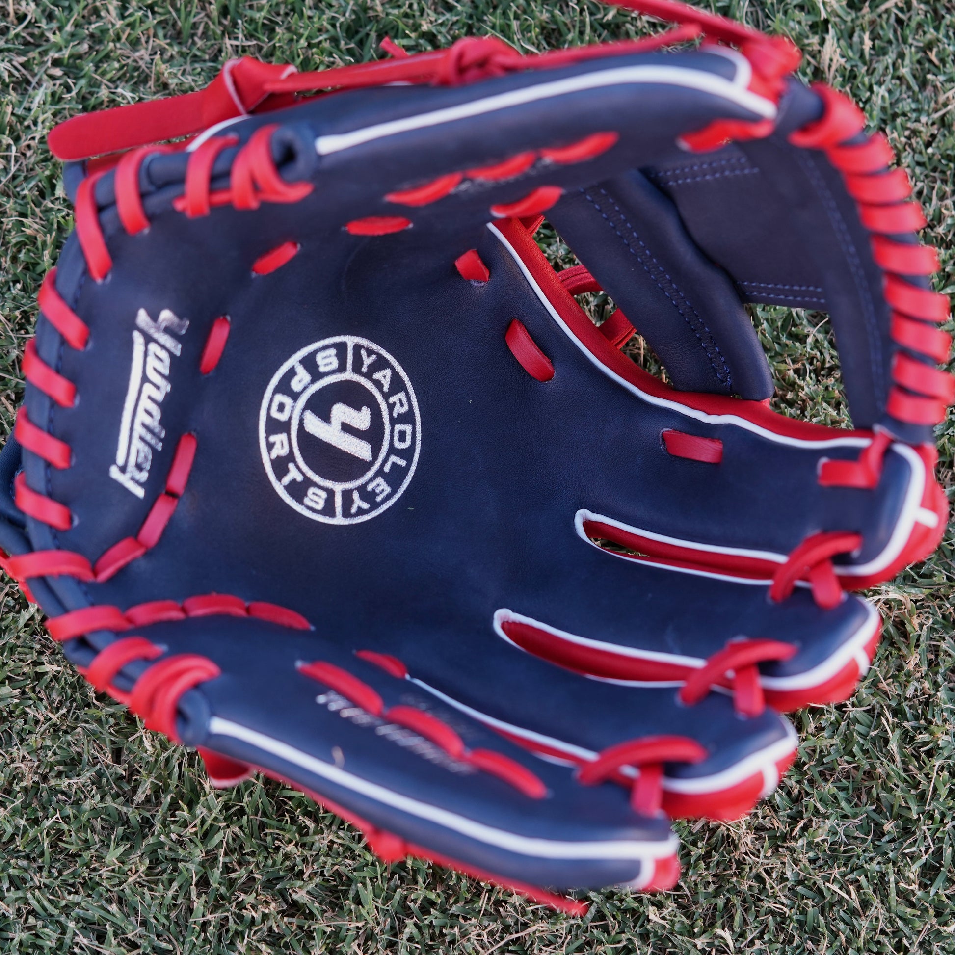 A red and navy blue baseball glove lying on grass, featuring the Yardley Sports logo and branding. The glove has red laces, white embroidered text, including the letter "Y." The design and color scheme highlight the premium quality and craftsmanship associated with Yardley Sports.