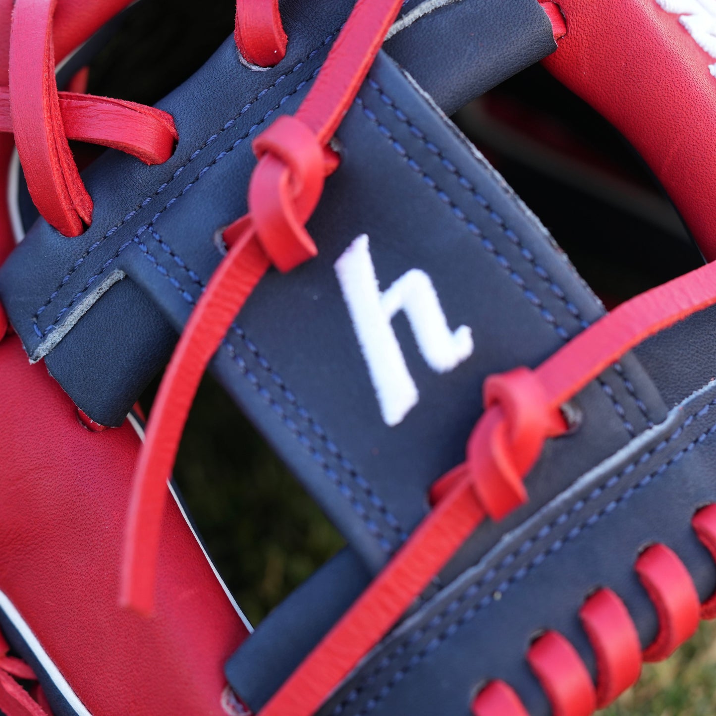 A red and navy blue baseball glove lying on grass, featuring the Yardley Sports logo and branding. The glove has red laces, white embroidered text, including the letter "Y." The design and color scheme highlight the premium quality and craftsmanship associated with Yardley Sports.