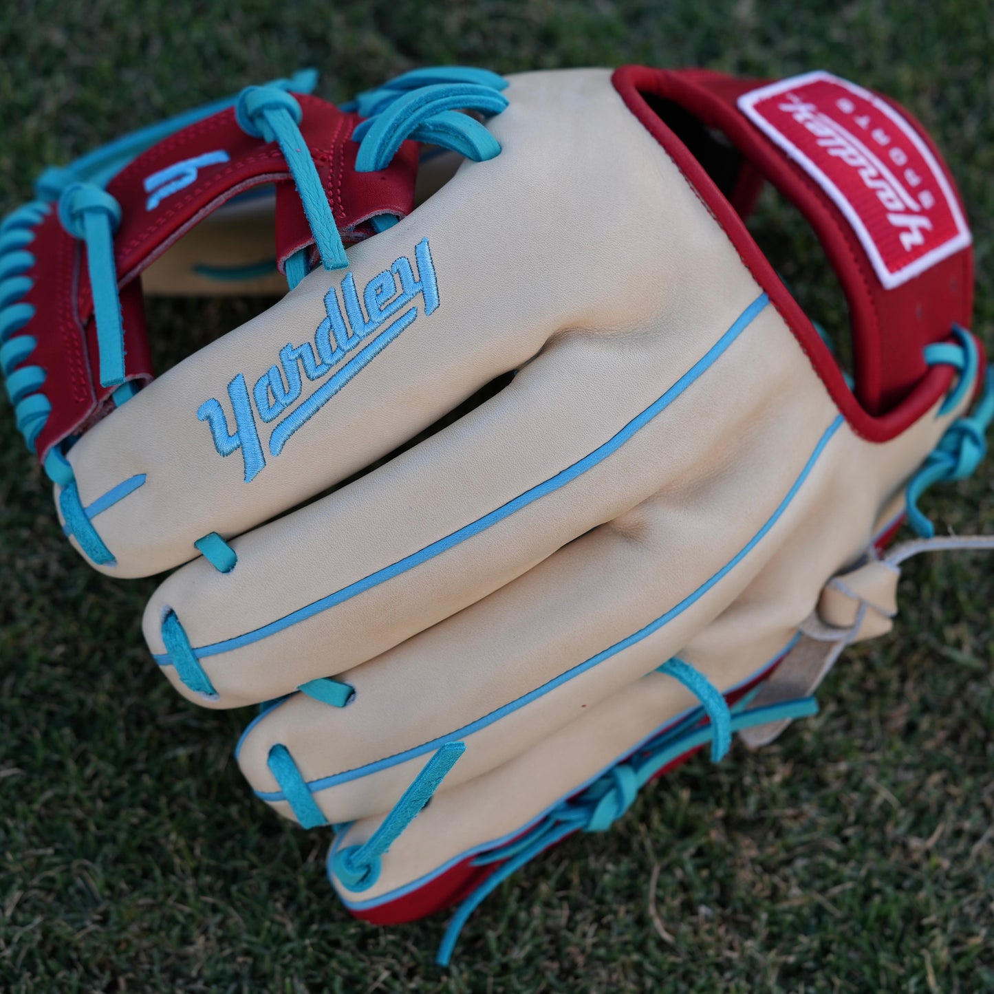 A stylish beige baseball glove with blue laces and accents, red trim around the wrist, and "Yardley" embroidered in blue on the back of the fingers. The glove rests on a grassy field, showcasing its vibrant design and quality craftsmanship.