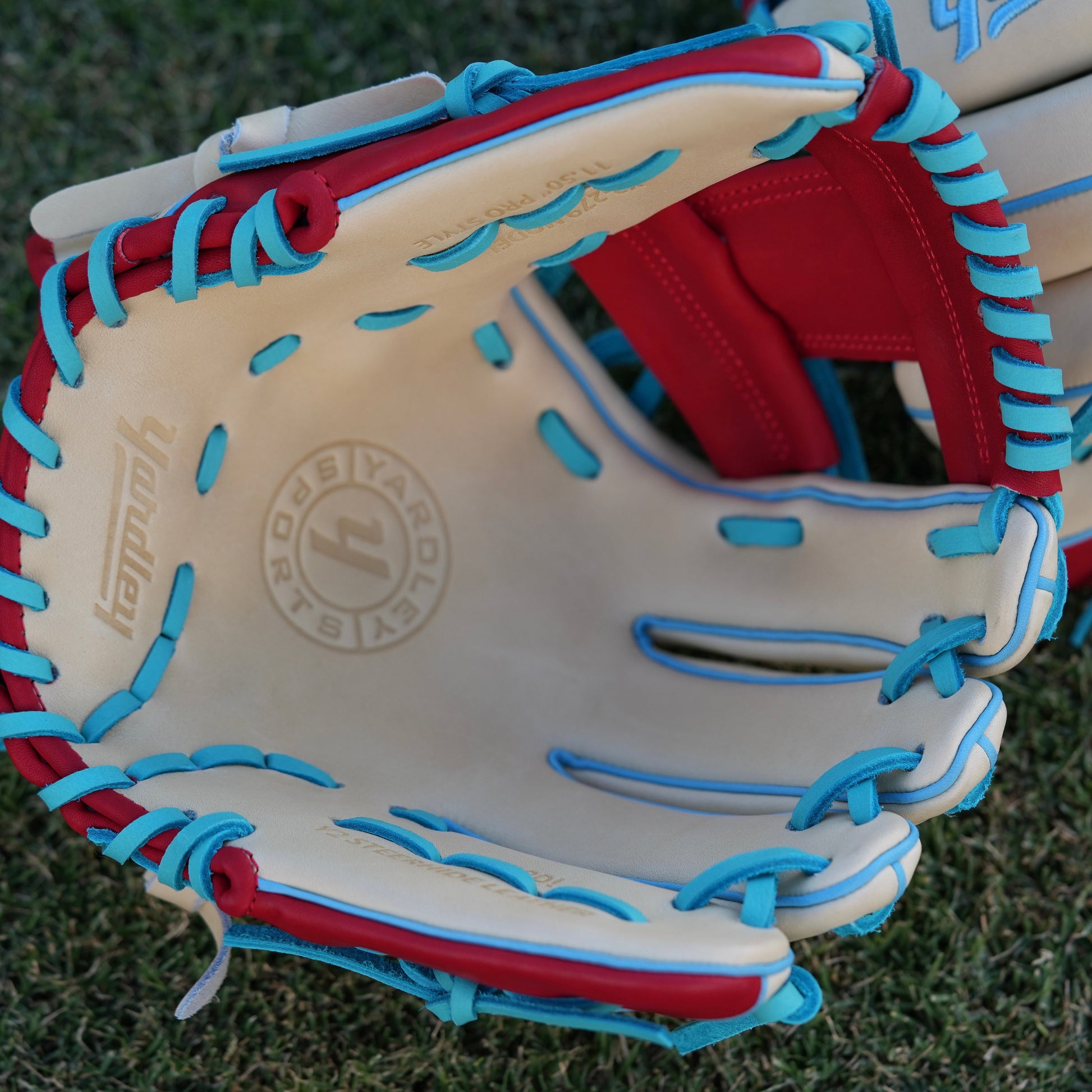 A stylish beige baseball glove with blue laces and accents, red trim around the wrist, and "Yardley" embroidered in blue on the back of the fingers. The glove rests on a grassy field, showcasing its vibrant design and quality craftsmanship.