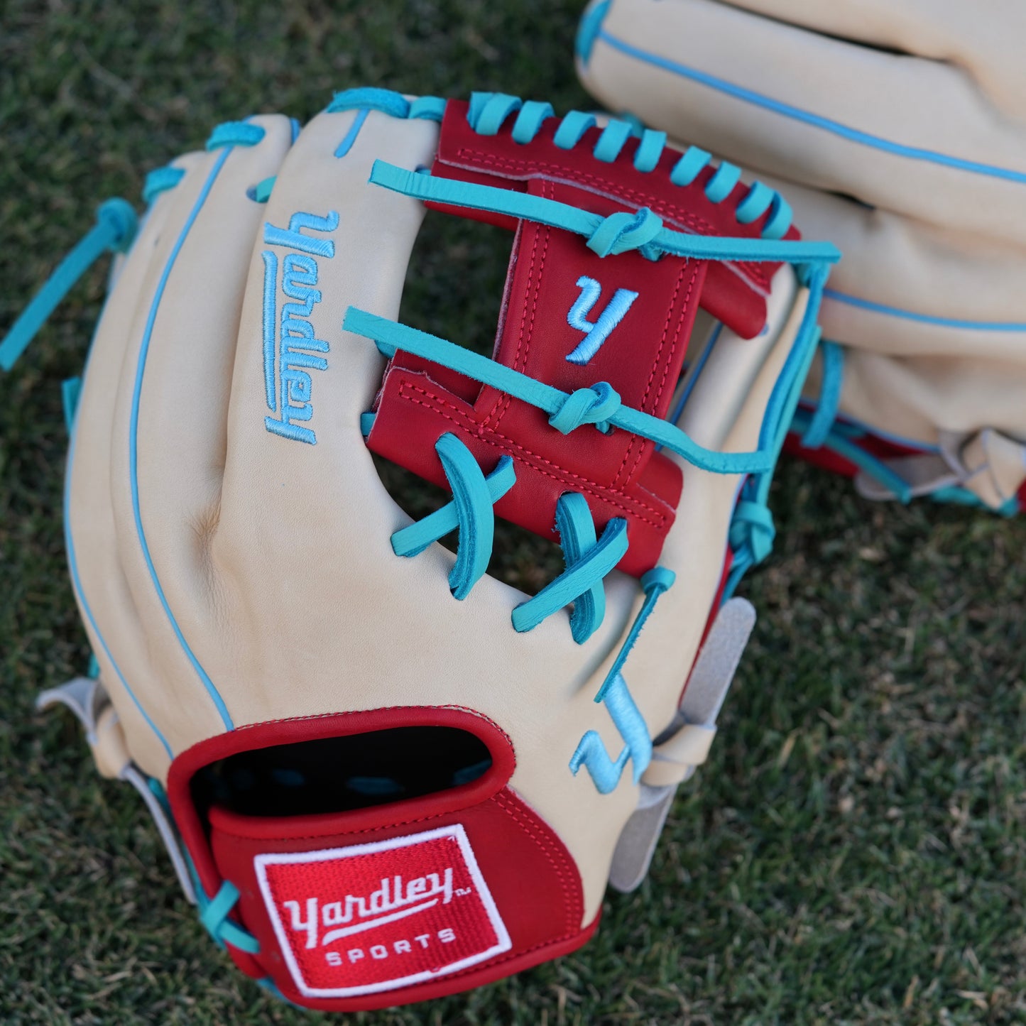 A stylish beige baseball glove with blue laces and accents, red trim around the wrist, and "Yardley" embroidered in blue on the back of the fingers. The glove rests on a grassy field, showcasing its vibrant design and quality craftsmanship.