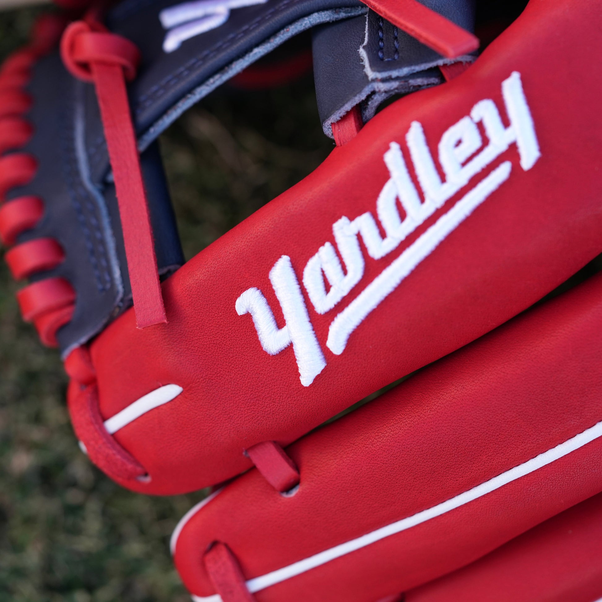 A red and navy blue baseball glove lying on grass, featuring the Yardley Sports logo and branding. The glove has red laces, white embroidered text, including the letter "Y." The design and color scheme highlight the premium quality and craftsmanship associated with Yardley Sports.