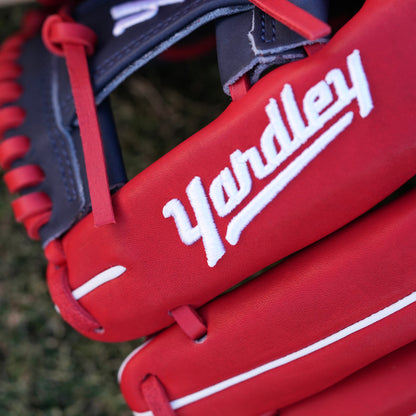 A red and navy blue baseball glove lying on grass, featuring the Yardley Sports logo and branding. The glove has red laces, white embroidered text, including the letter "Y." The design and color scheme highlight the premium quality and craftsmanship associated with Yardley Sports.