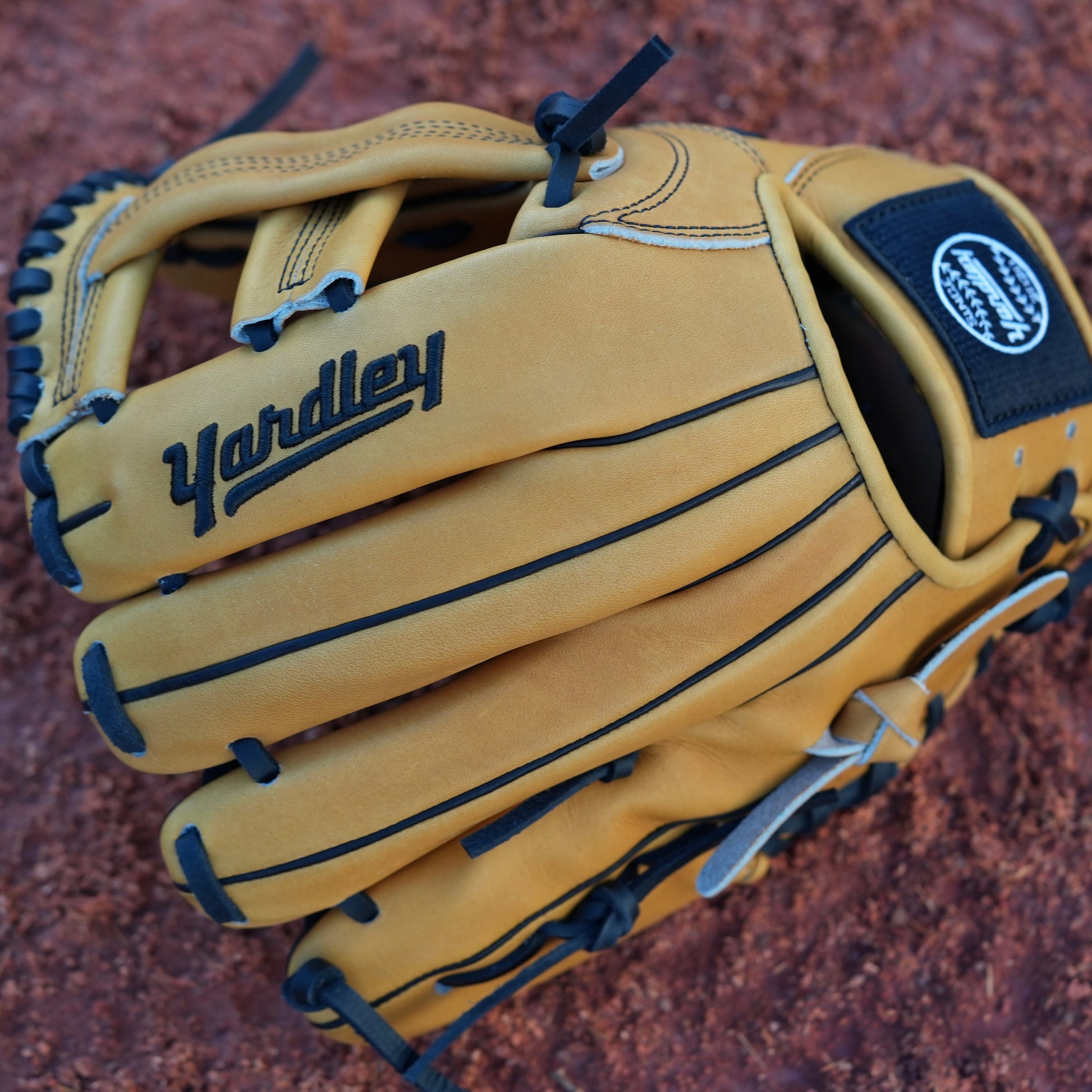 Tan baseball glove with black laces, resting on a dirt baseball field. The brand name "Yardley" is prominently displayed in black lettering.