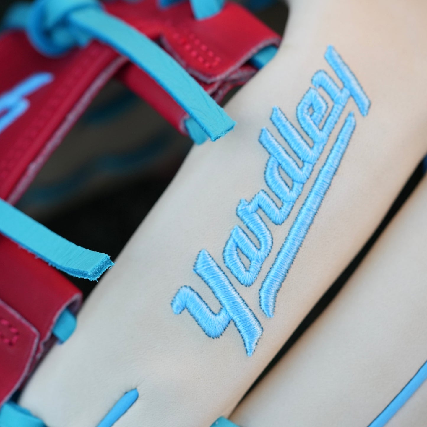 A stylish beige baseball glove with blue laces and accents, red trim around the wrist, and "Yardley" embroidered in blue on the back of the fingers. The glove rests on a grassy field, showcasing its vibrant design and quality craftsmanship.