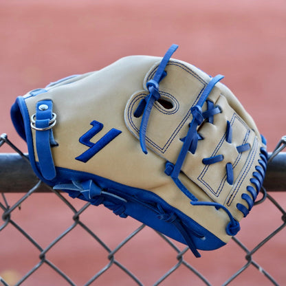 A tan, closed-back baseball glove with blue laces and the letter "Y" stitched in blue, hanging on a chain-link fence. The blurred background shows a red dirt baseball field.