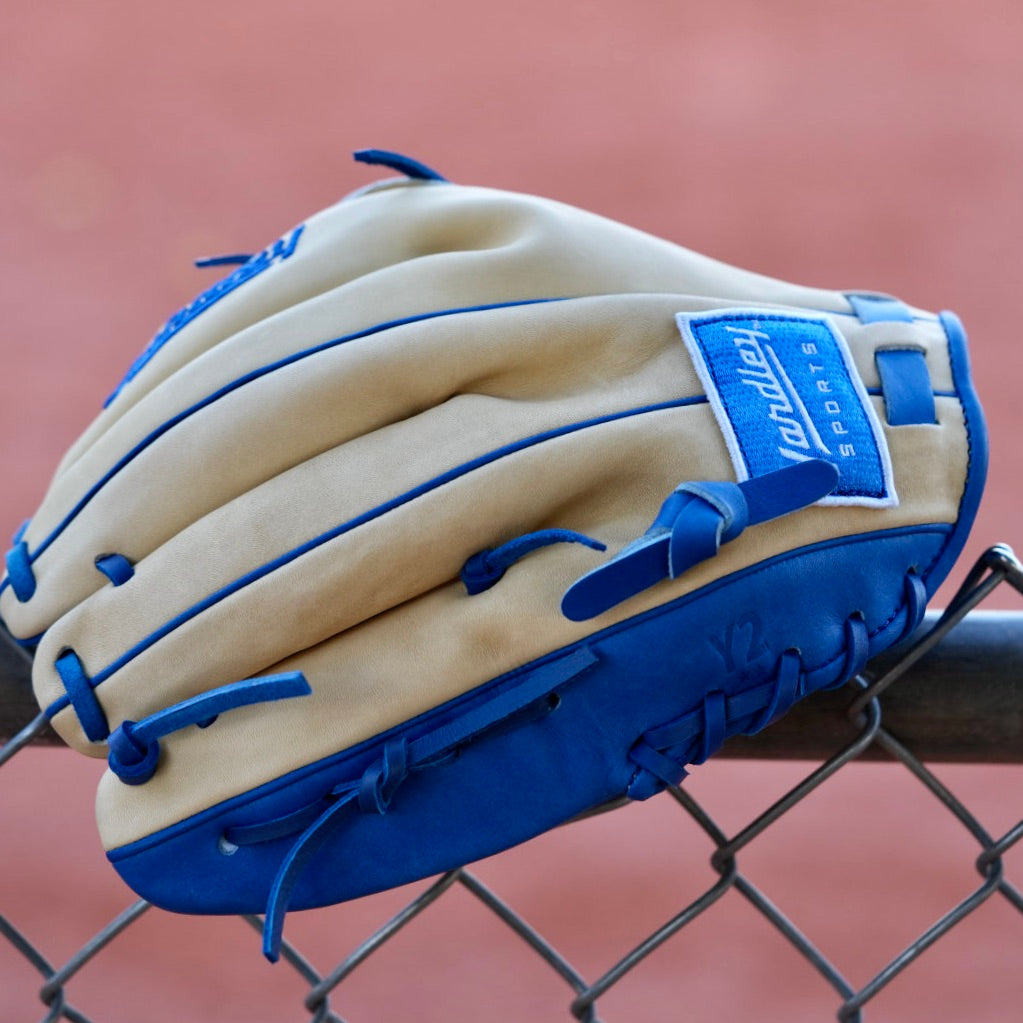 A tan, closed-back baseball glove with blue laces and the letter "Y" stitched in blue, hanging on a chain-link fence. The blurred background shows a red dirt baseball field.