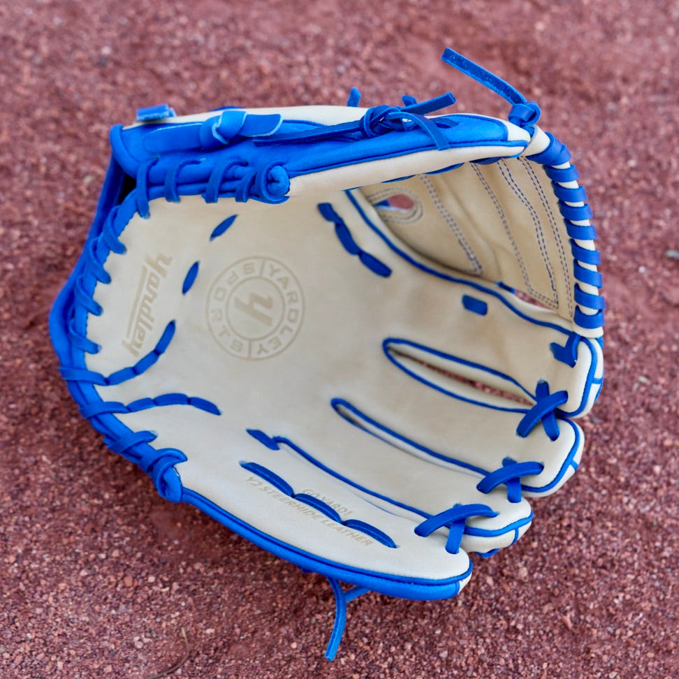 A tan, closed-back baseball glove with blue laces and the letter "Y" stitched in blue, hanging on a chain-link fence. The blurred background shows a red dirt baseball field.