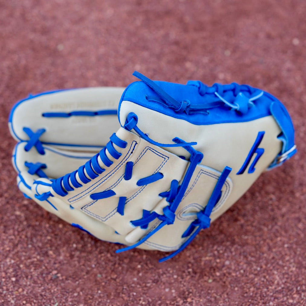 A tan, closed-back baseball glove with blue laces and the letter "Y" stitched in blue, hanging on a chain-link fence. The blurred background shows a red dirt baseball field.