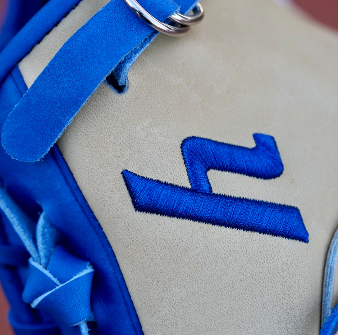 A tan, closed-back baseball glove with blue laces and the letter "Y" stitched in blue, hanging on a chain-link fence. The blurred background shows a red dirt baseball field.