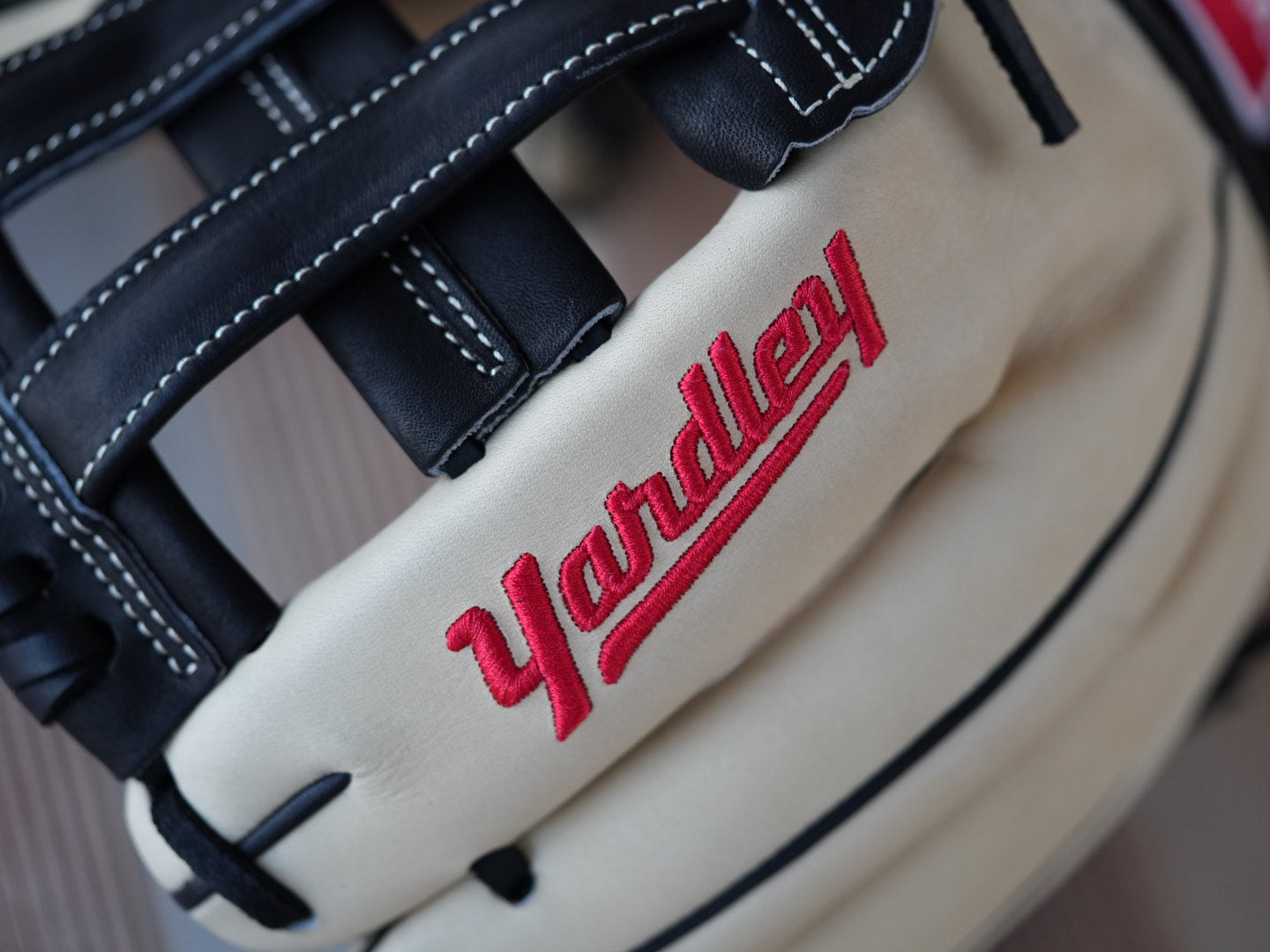 Beige baseball glove with black stitching and red "Yardley" branding, placed on a metal surface. The glove showcases a sleek design with fine craftsmanship.