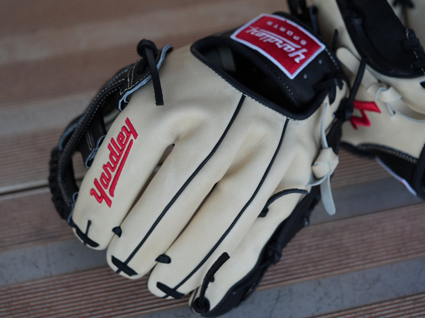 Beige baseball glove with black stitching and red "Yardley" branding, placed on a metal surface. The glove showcases a sleek design with fine craftsmanship.