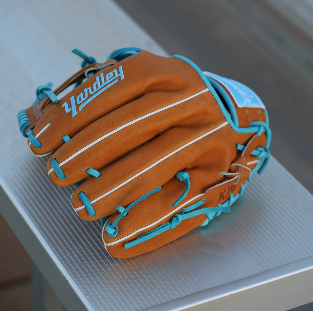 A brown and blue Yardley baseball glove with blue laces and stitching sits on a metallic surface. The glove features the letter Y stitched in blue on the thumb area and another letter Y on the webbing. The modern, stylish design showcases a unique color combination and intricate craftsmanship.