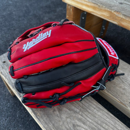  A red Yardley baseball glove with black and white stitching and black laces on a wooden bench. The glove has a "Y" logo on the thumb area, showcasing its unique design. The webbing is an H-web style