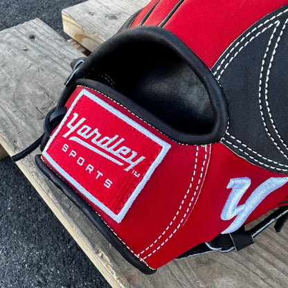  A red Yardley baseball glove with black and white stitching and black laces on a wooden bench. The glove has a "Y" logo on the thumb area, showcasing its unique design. The webbing is an H-web style