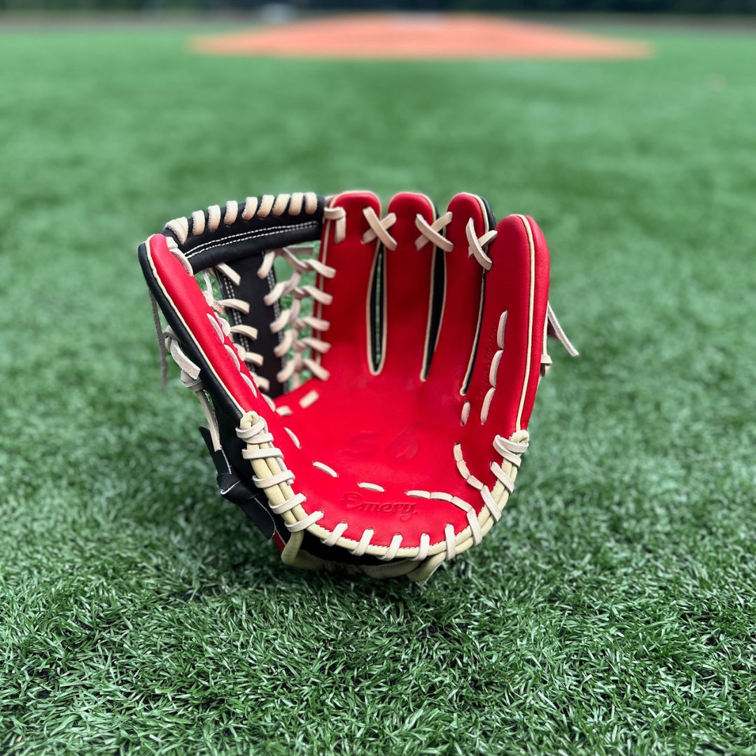 A close-up image of a red and black baseball glove resting on a grassy field. The glove is positioned with the palm facing forward, showcasing its intricate blonde lacing and padded leather design with a striking red palm. In the background, there is a blurred view of a baseball diamond, indicating that the setting is likely a sports field. The focus on the glove against the soft-focus backdrop highlights its details and suggests its readiness for use in a game.