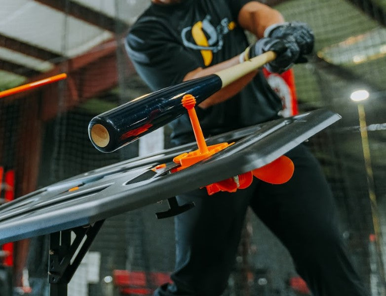 A baseball batter taking a swing inside an indoor baseball training facility. A wood bat is about to connect with the dry swing tee topper (orange)